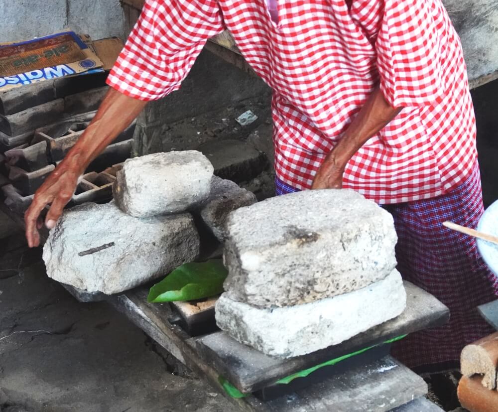 The mold and its contents are covered with a fresh green palm leave and a wooden plank and heavy stones placed on top
